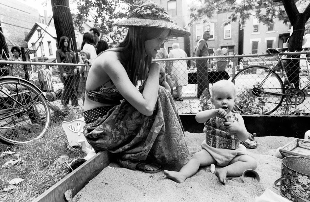A woman in a sunhat sits beside a baby playing in a sandbox at a park. The scene is outdoors with people, bikes, and houses in the background, suggesting a community gathering. The image is in black and white.