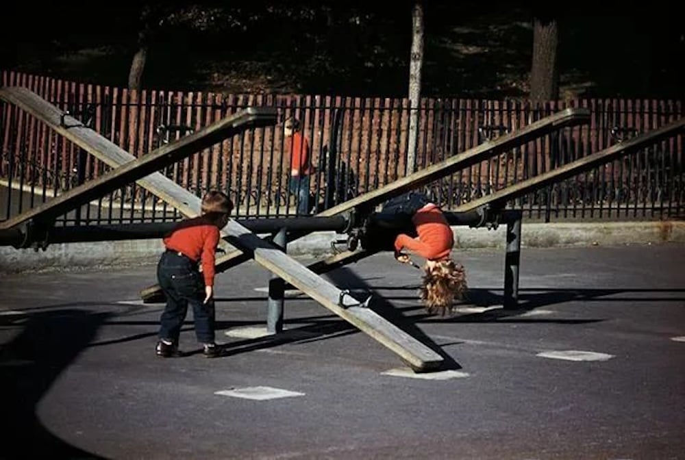 Two children in orange shirts play on old wooden seesaws in a park. One child is upside down on a seesaw, while the other stands nearby. A third child is visible in the background near a fence.