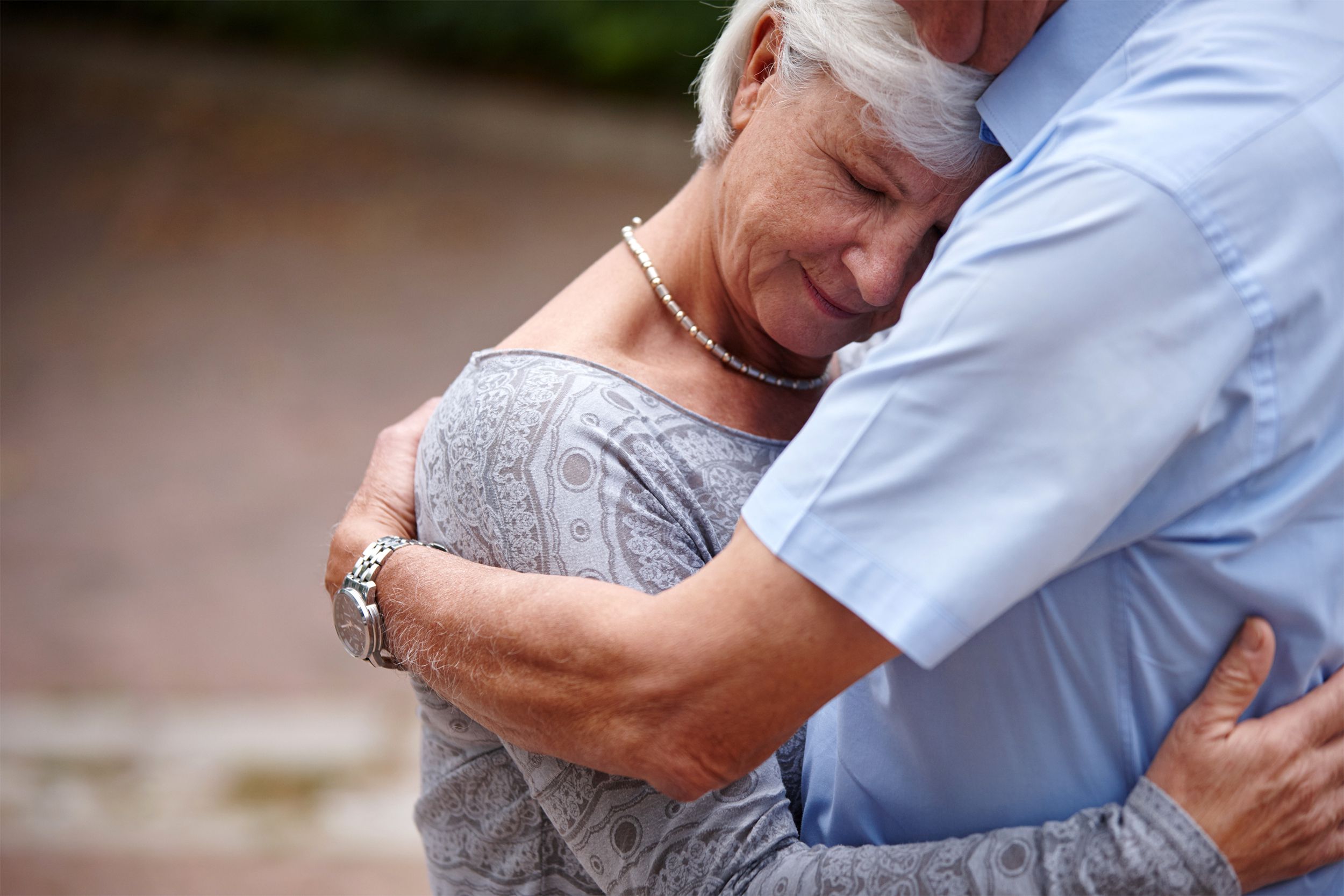Senior woman hugging her overweight senior man, she is resting her head on his chest, happy, a blurred background of beige on the left