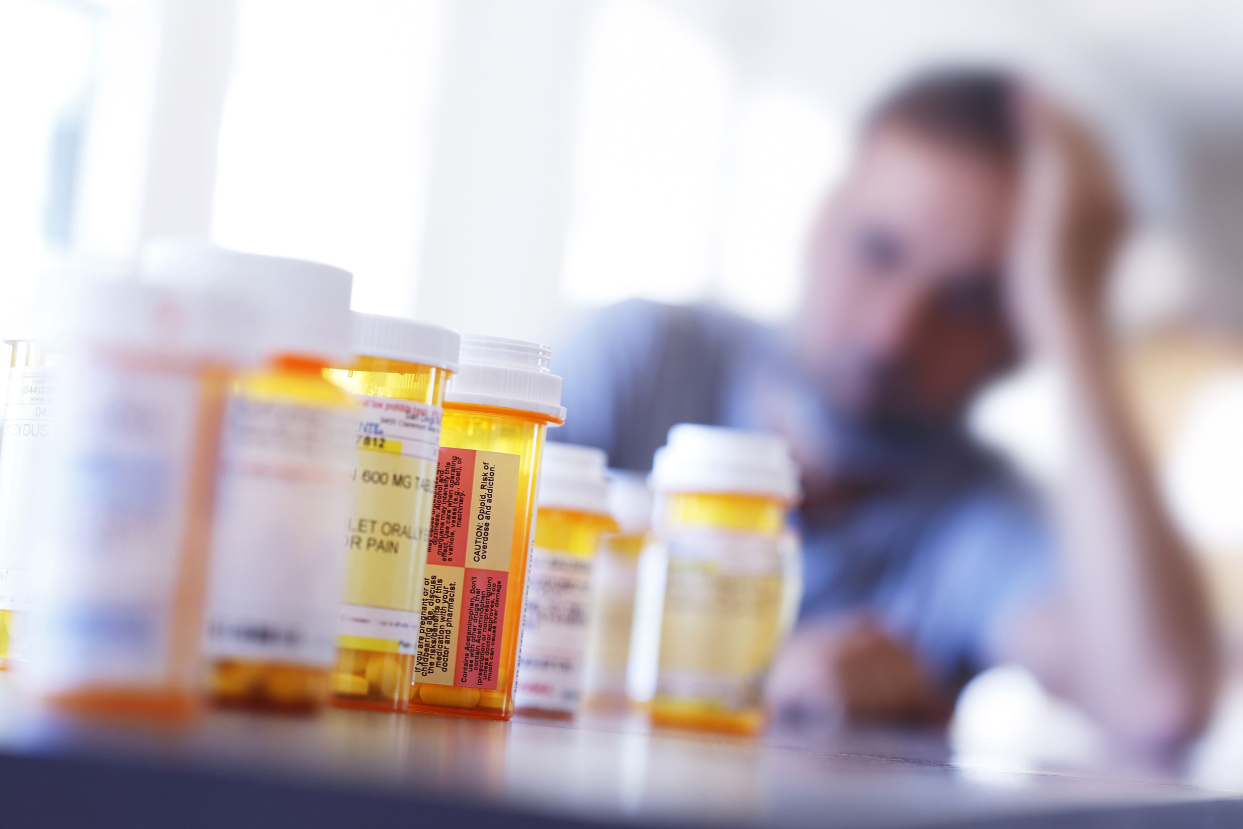 Several yellow prescription bottles on a table in the foreground, center in focus, surrounding bottles blurred, a man with his hand holding his head facing the bottle, blurred in the background with a wall of windows sunlight coming in