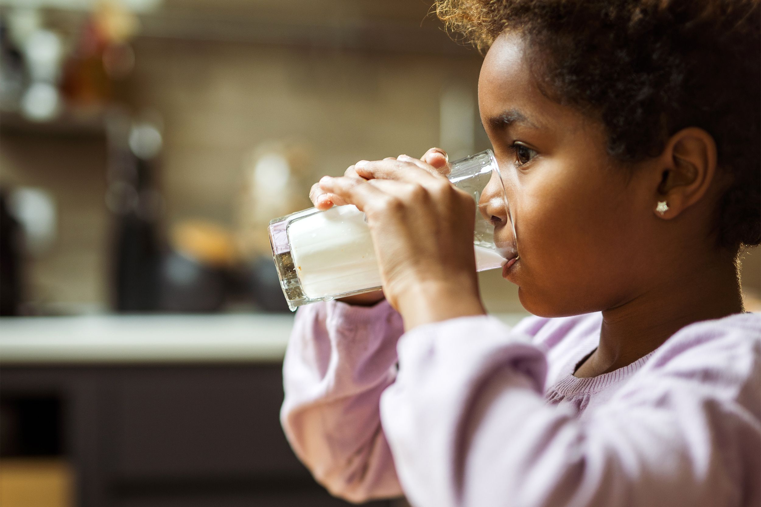 Sideview of a young girl drinking a glass of milk, selective focus of her on right side with a blurred background of her kitchen