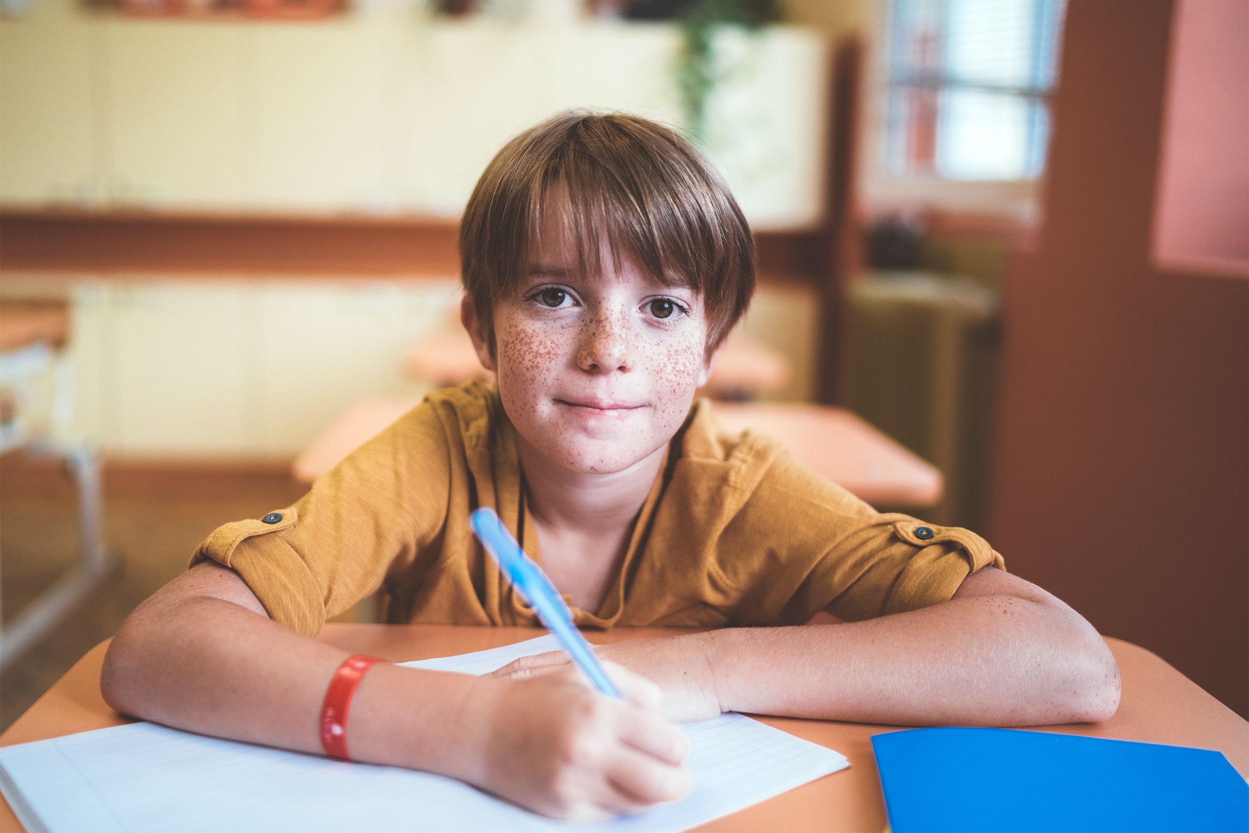 Smiling boy with freckles sitting at his desk in school, writing in a notebook with a pen in the right hand, looking into the camera, with a blurred background of an empty back of classroom