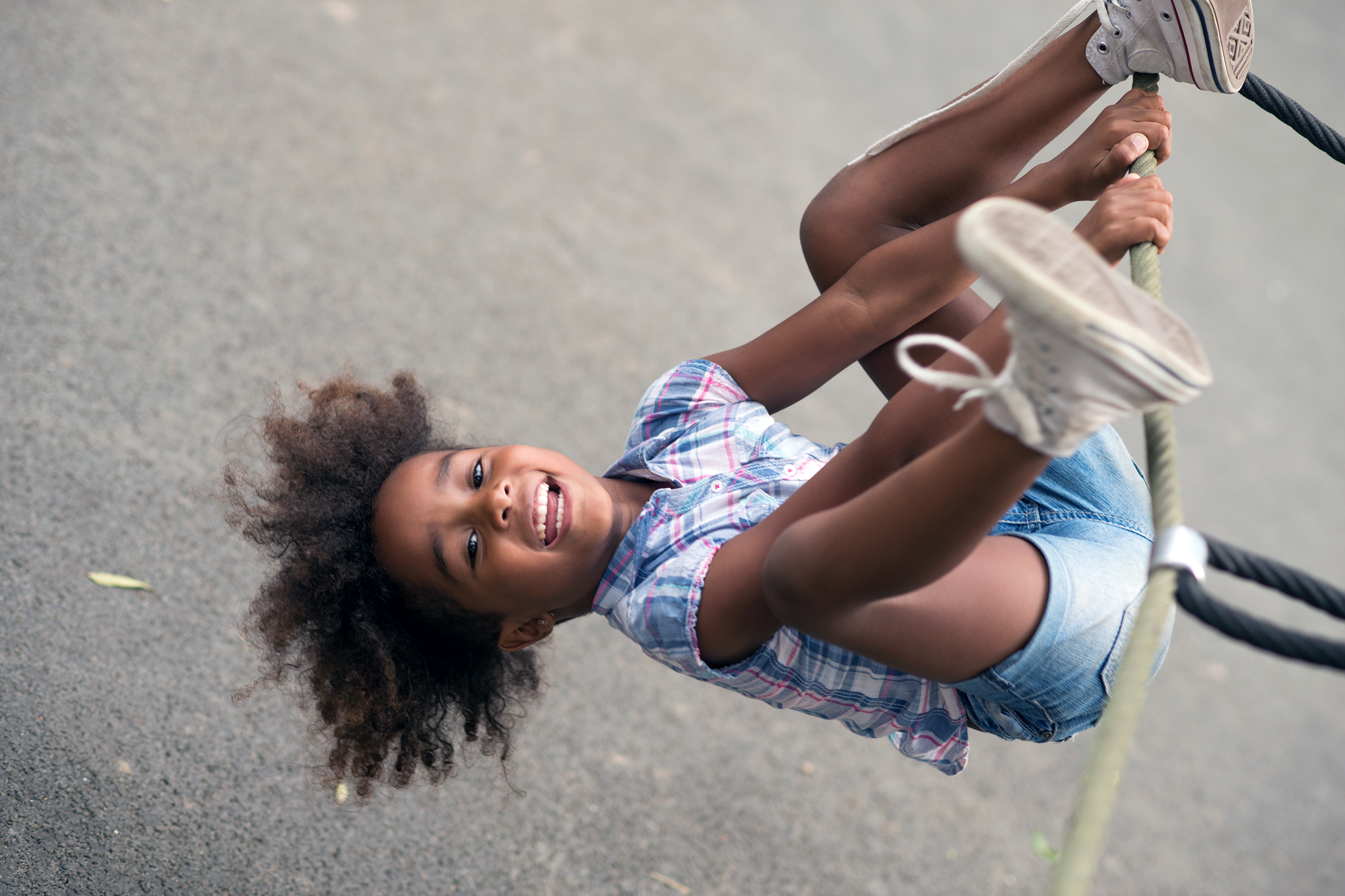 Smiling little girl hanging upside down from the side of a rope jungle gym over concrete, in a playground
