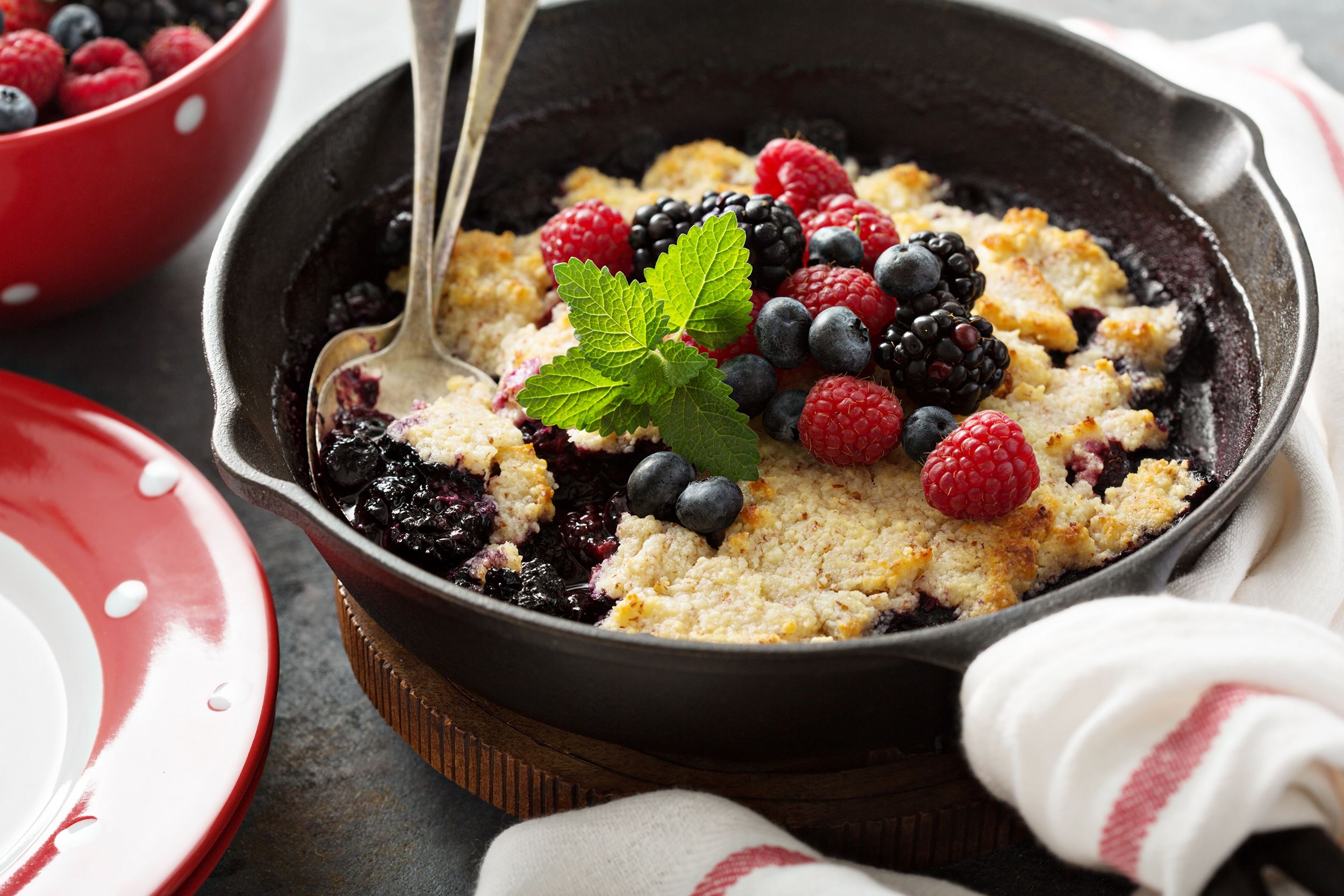 Sugar-free low carb blackberry cobbler in an iron skillet with two spoons surrounded by a red and white polka dot plate, a bowl of berries, and a matching napkin