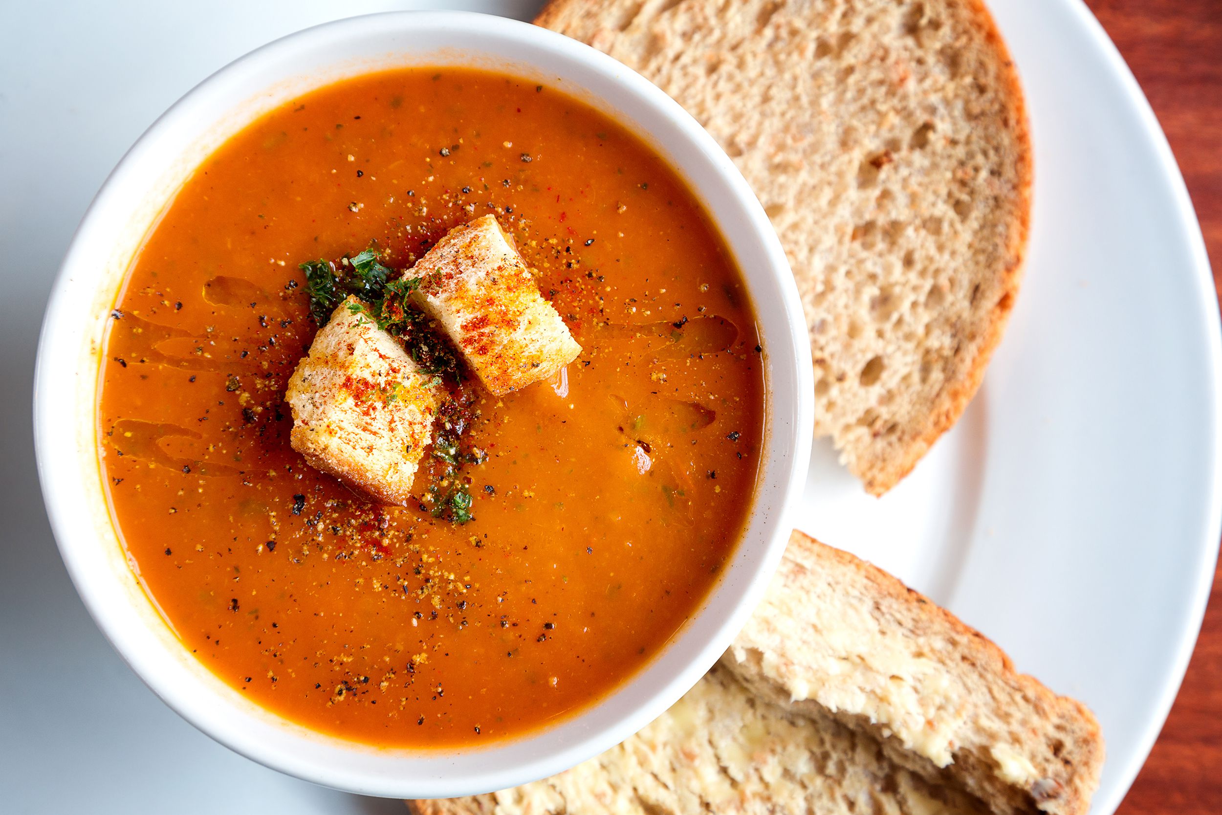 Top-view of no-cook blender tomato soup in a white porcelain bowl with two large croutons, on the left with two pieces of bread, all on a white porcelain plate, on a table, shown on both top and bottom right