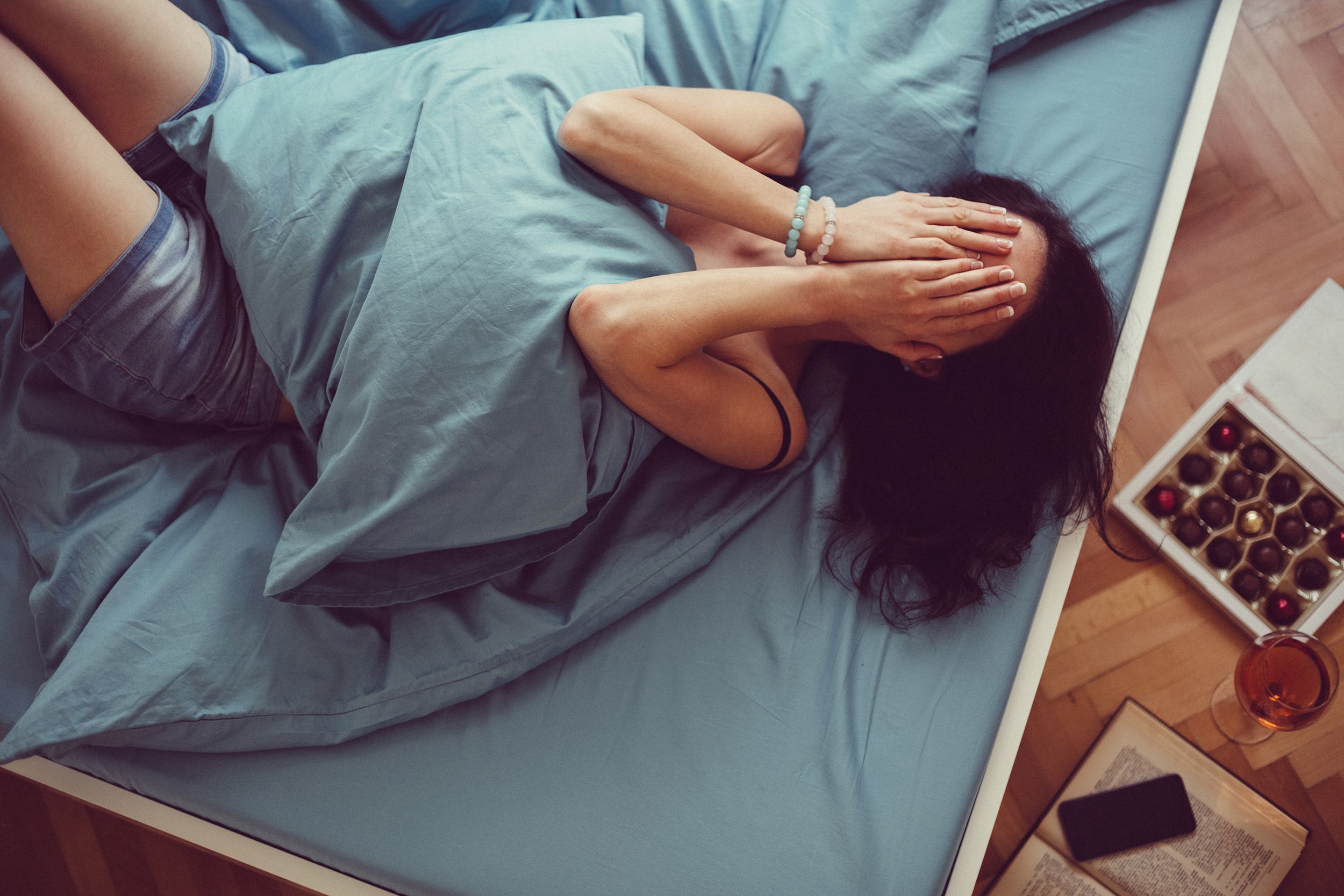 Top-view of a woman laying on her bed facing up with her hands covering her eyes, steel-grey covers, sheets, and pillow, a box of chocolates, a glass of rose wine, a smartphone on a book, on the floor on the right