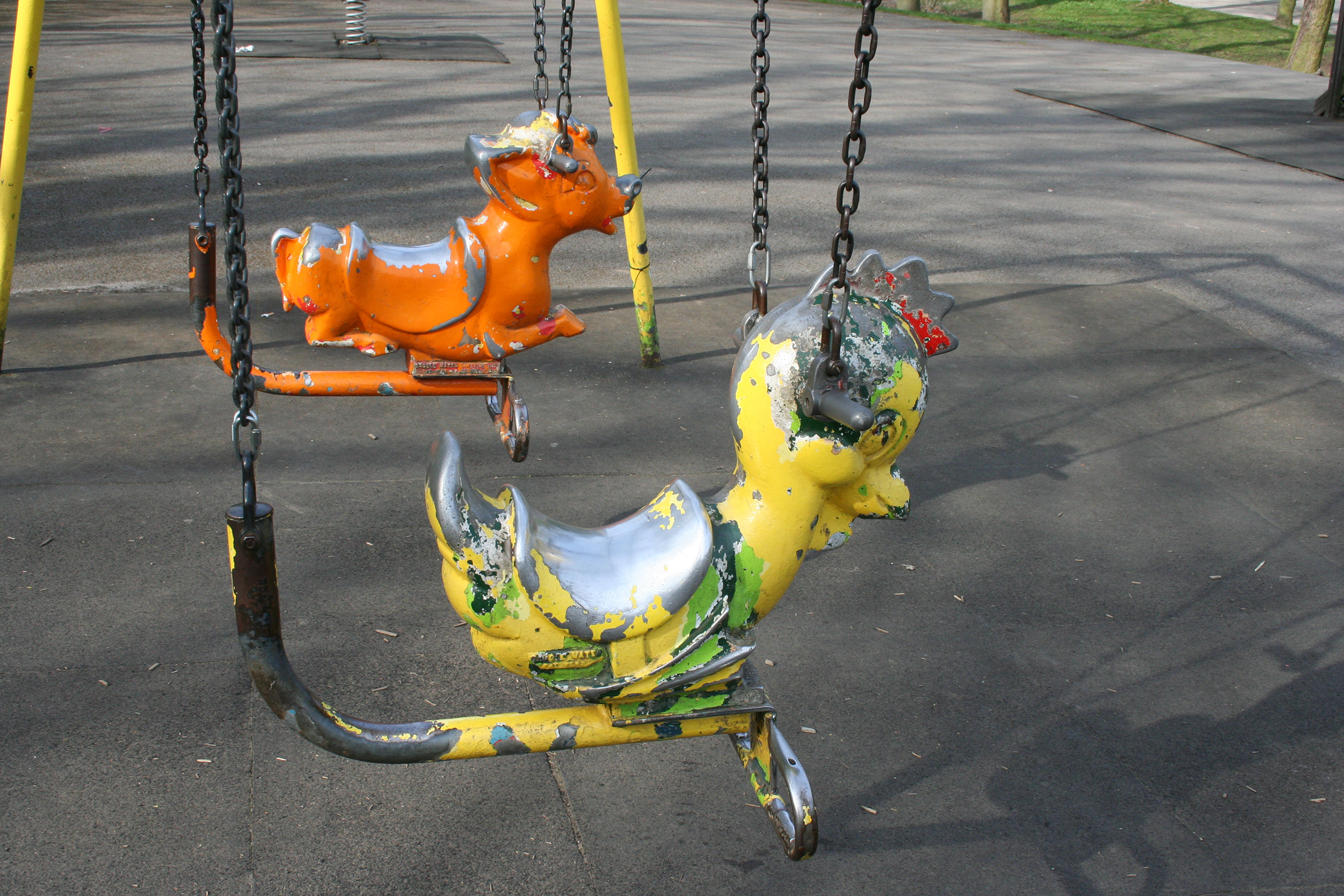 Two metal animal swings, a yellow chicken in the foreground with an orange cow behind it, both with worn paint, in a playground on cement ground