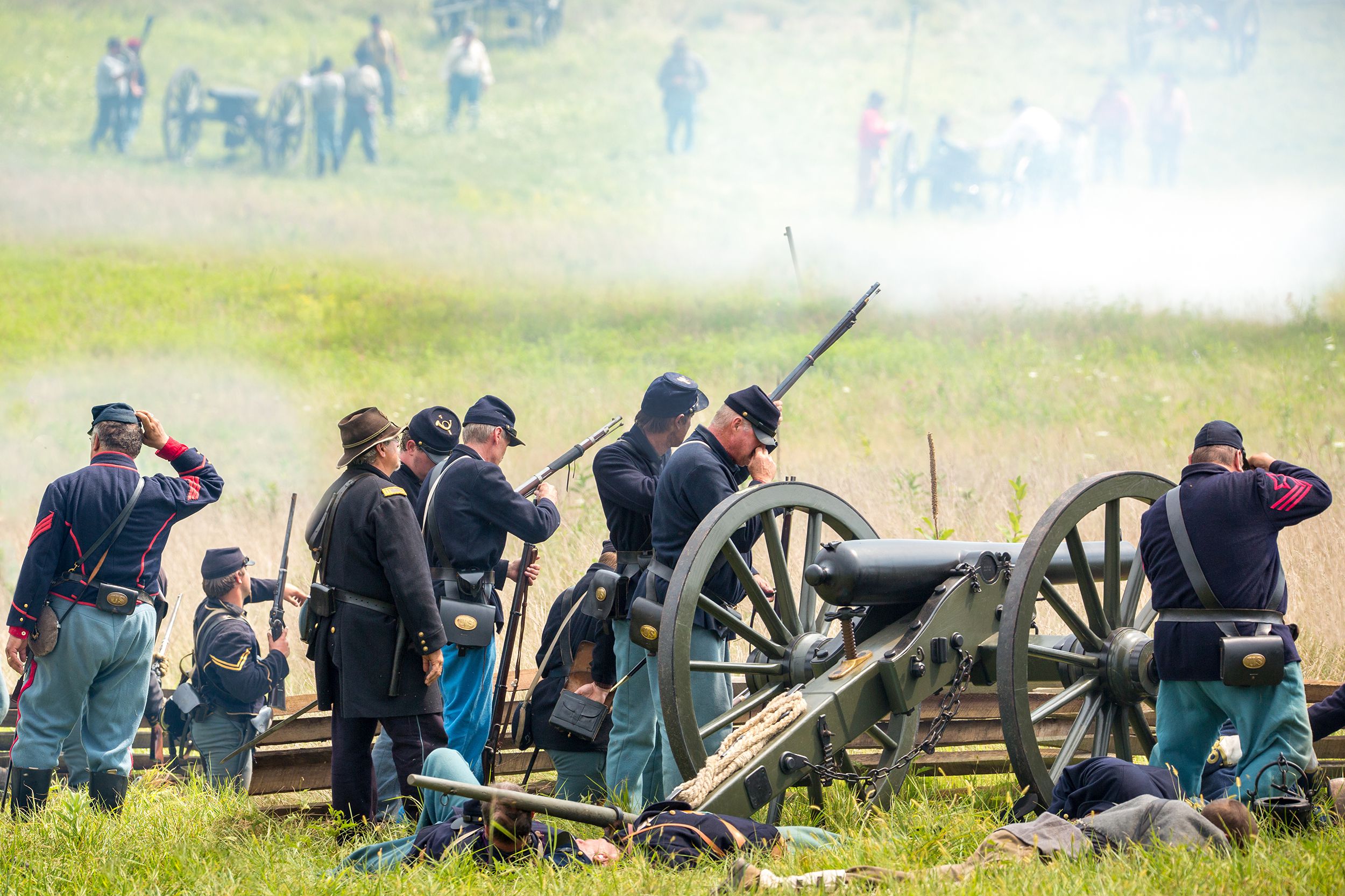 Union soldiers fight during a reenactment of the Civil War battle of Gettysburg, Pennsylvania, soldiers with a cannon projectile and wooden fence on a grassy hill in the foreground, with more soldiers and field artillery in dust in the background