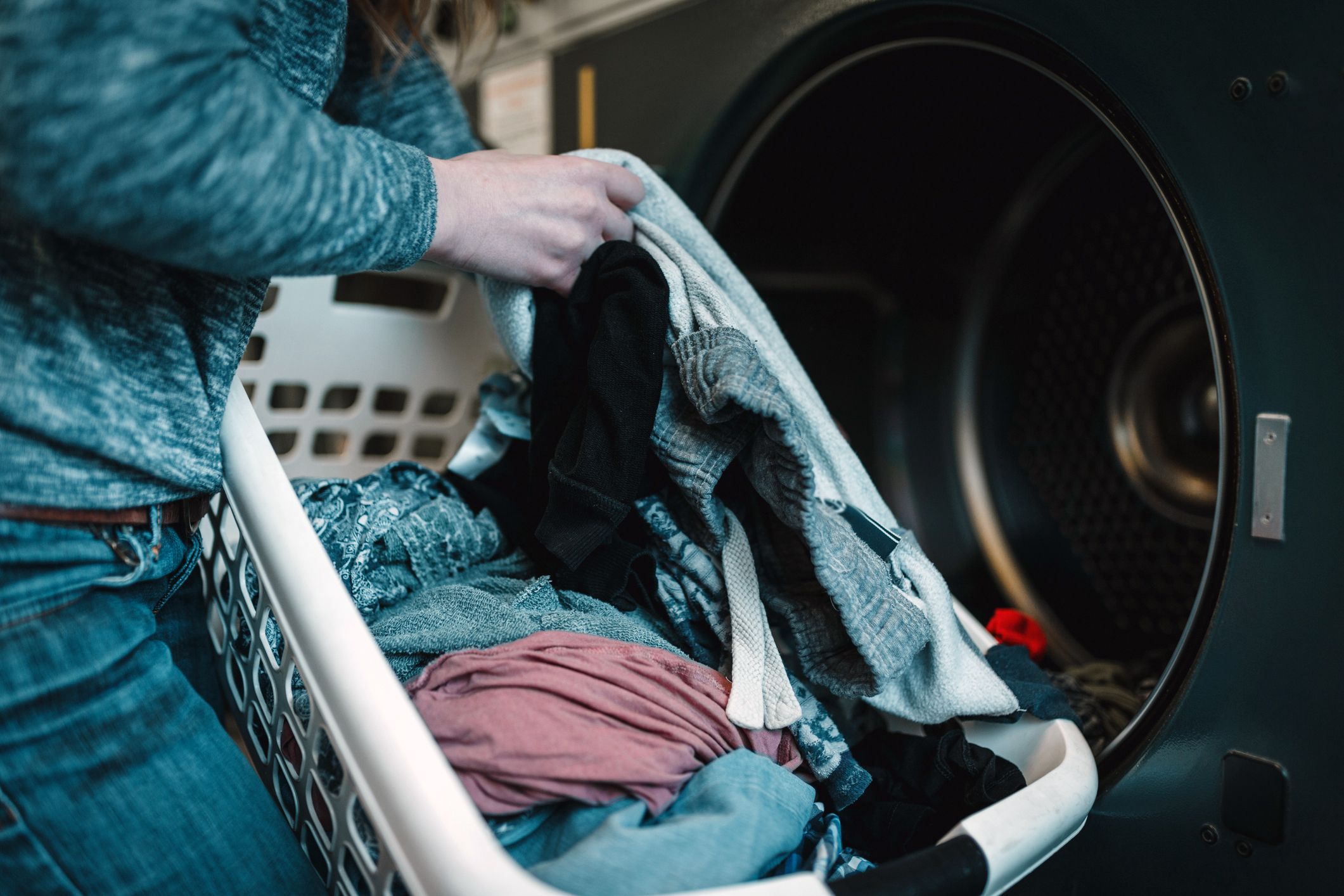 Woman pulling clean clothes out of dryer, focus on white laundry basket in front of it, opened grey dryer in the background