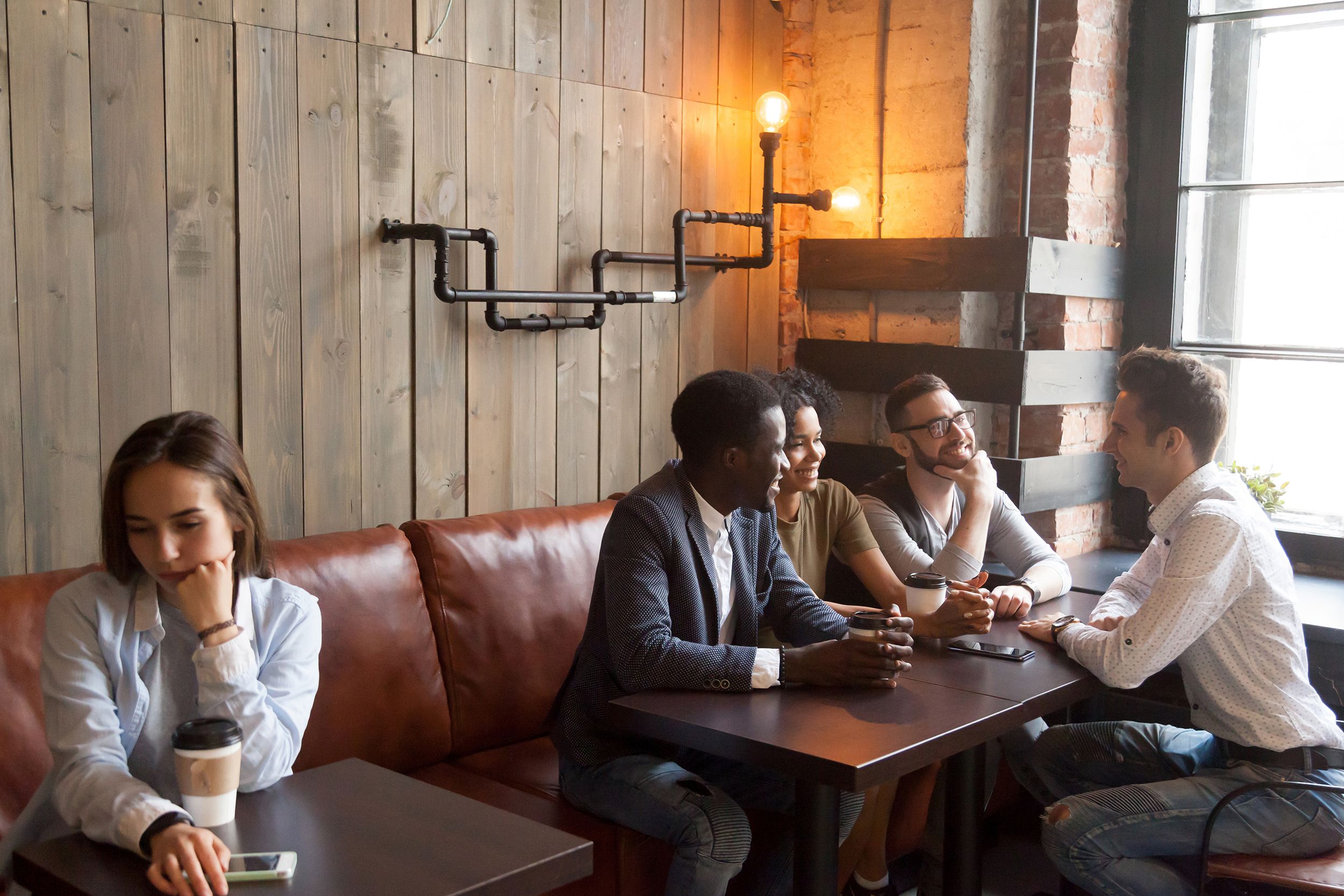 Woman sitting alone at a small table in a coffee shop on the left, a group of four friends sitting at two adjacent tables on the right, rustic pipe light on a wooden wall in the background, hipster vibes