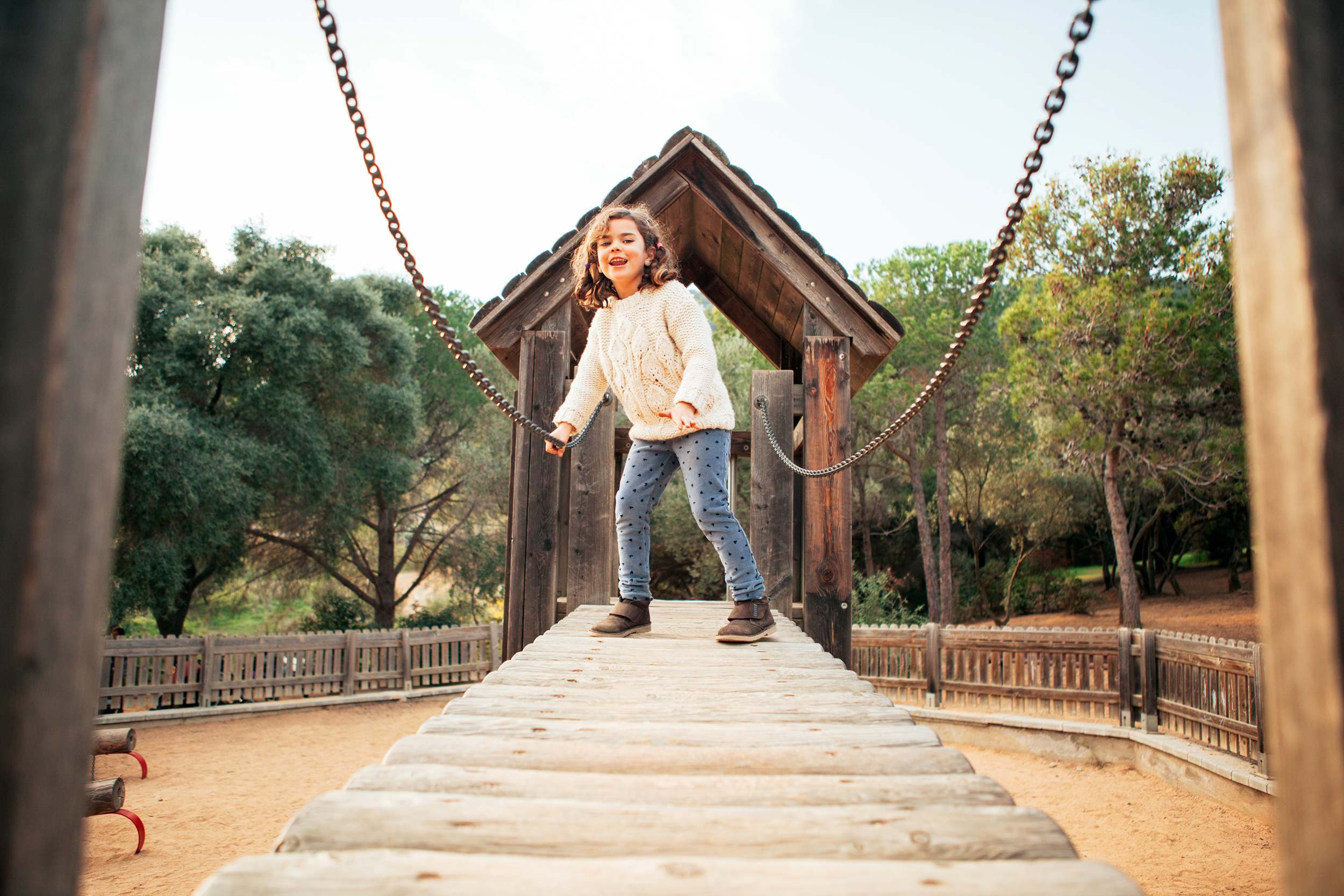 Young girl walking across a wooden bridge, as seen from the other side of it, she is holding onto one metal chain rail, on sand in a playground, with trees in the background in autumn