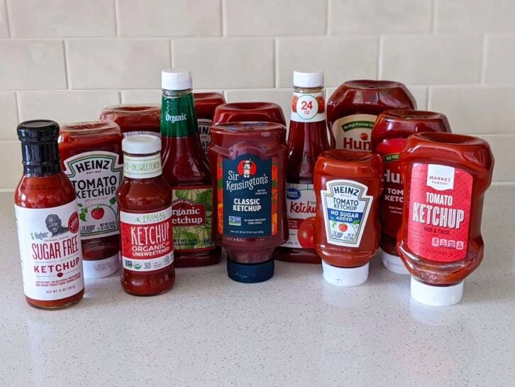 A variety of ketchup bottles from different brands, including Heinz, Hunt’s, Sir Kensington’s, and store brands, are arranged on a white countertop with a tiled background.