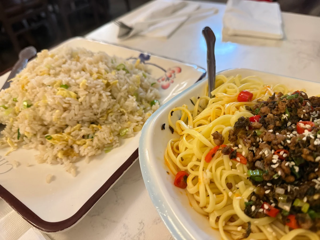A plate of fried rice with vegetables next to a bowl of noodles topped with minced meat, sliced red chili, green onions, and sesame seeds on a white table.