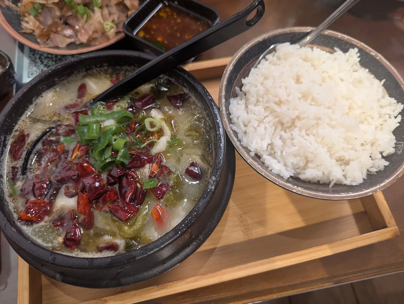 A bowl of spicy soup topped with chopped green onions and red chili peppers sits next to a bowl of steamed white rice on a wooden tray. A spoon is in each bowl.