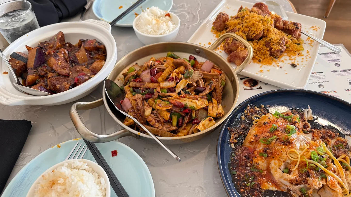 Four plates of Chinese-style dishes on a table, including stir-fried vegetables with noodles, braised eggplant, battered fried pieces, and a baked seafood dish, with two bowls of white rice nearby.
