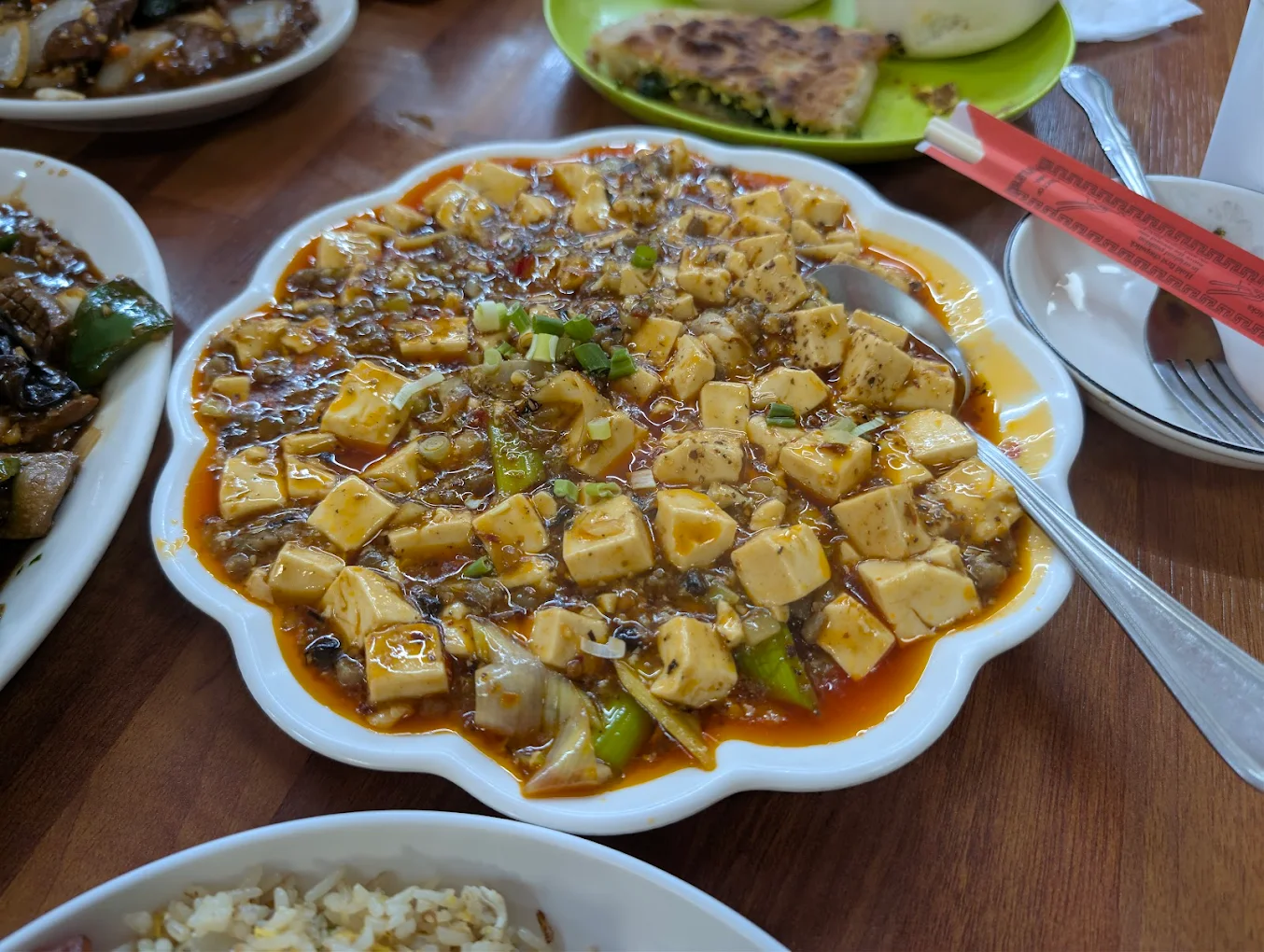 A plate of mapo tofu with cubed tofu, ground meat, and green onions in a spicy red sauce, surrounded by other Chinese dishes on a wooden table. A spoon rests in the dish.