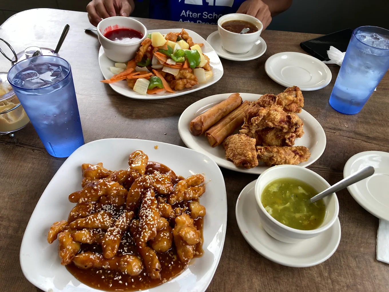 A table with plates of Chinese food, including sesame chicken, fried chicken wings with egg rolls, sweet and sour chicken with vegetables, a bowl of egg drop soup, and two glasses of water.