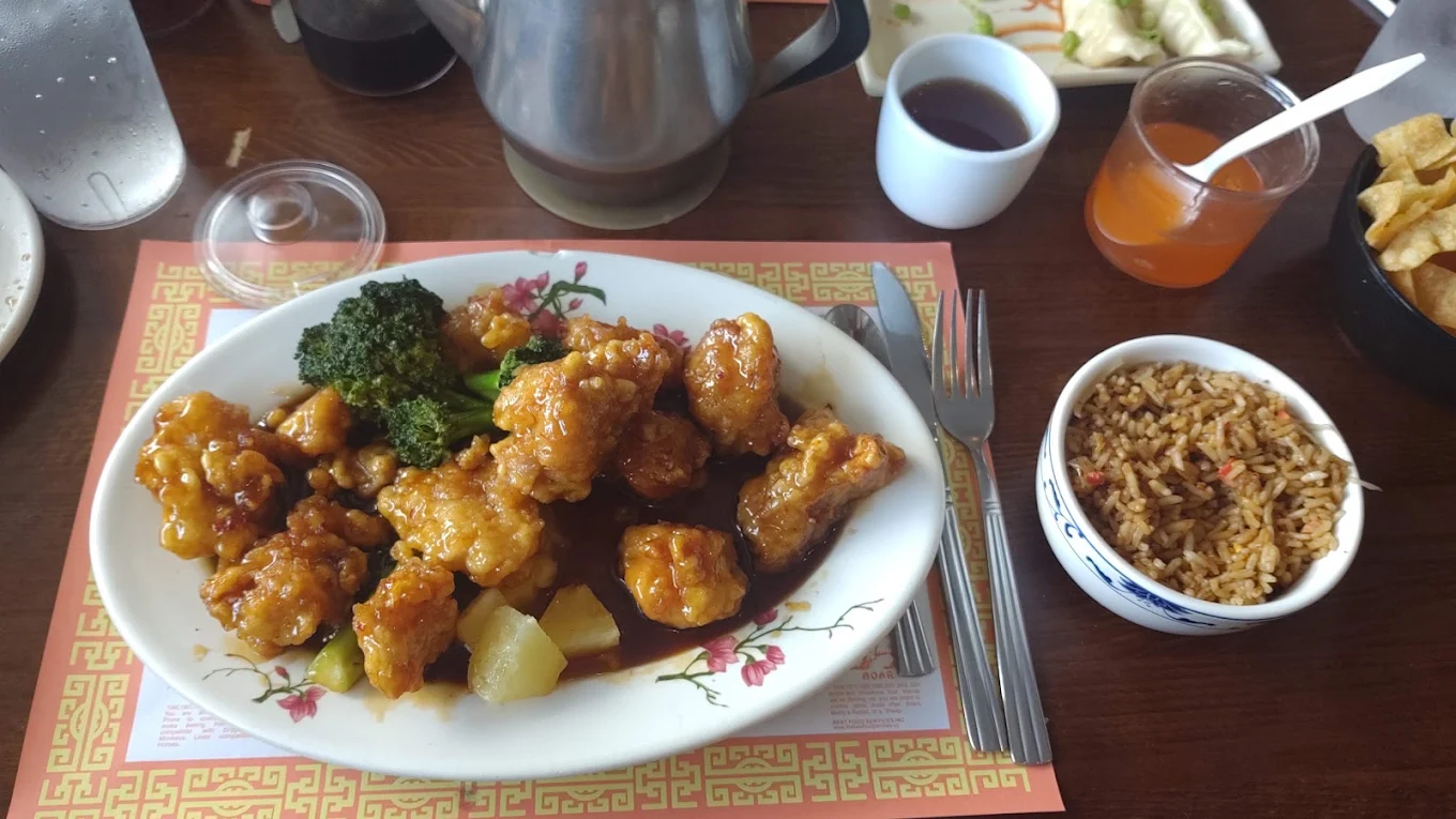 A plate of sweet and sour chicken with broccoli and pineapple, next to a bowl of fried rice. Drinks, sauces, and other side dishes are also on the table.