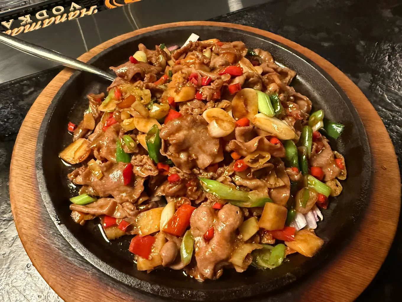 A sizzling plate of stir-fried beef with bell peppers, onions, sliced garlic, and green onions in a savory sauce, served on a wooden platter.