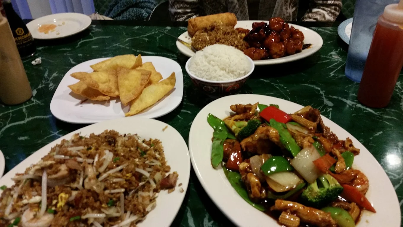 A table with various Chinese dishes, including fried rice, stir-fried vegetables with chicken, crab rangoons, egg roll, white rice, and a plate of sweet and sour chicken.