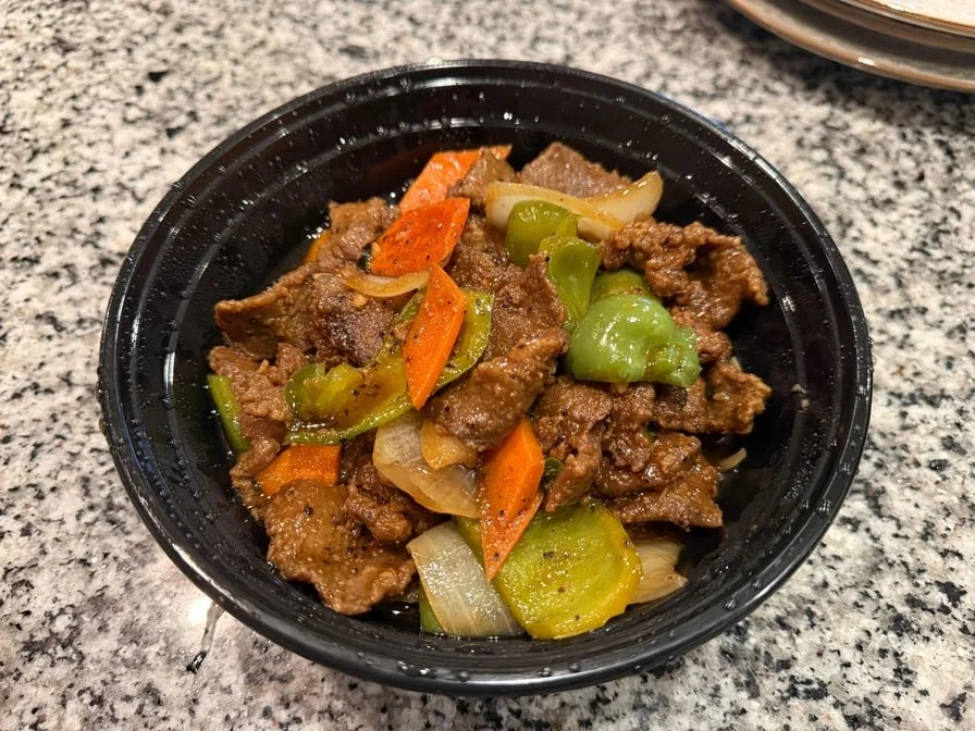 A black bowl filled with stir-fried beef, sliced carrots, green bell peppers, and onions, placed on a speckled granite countertop.