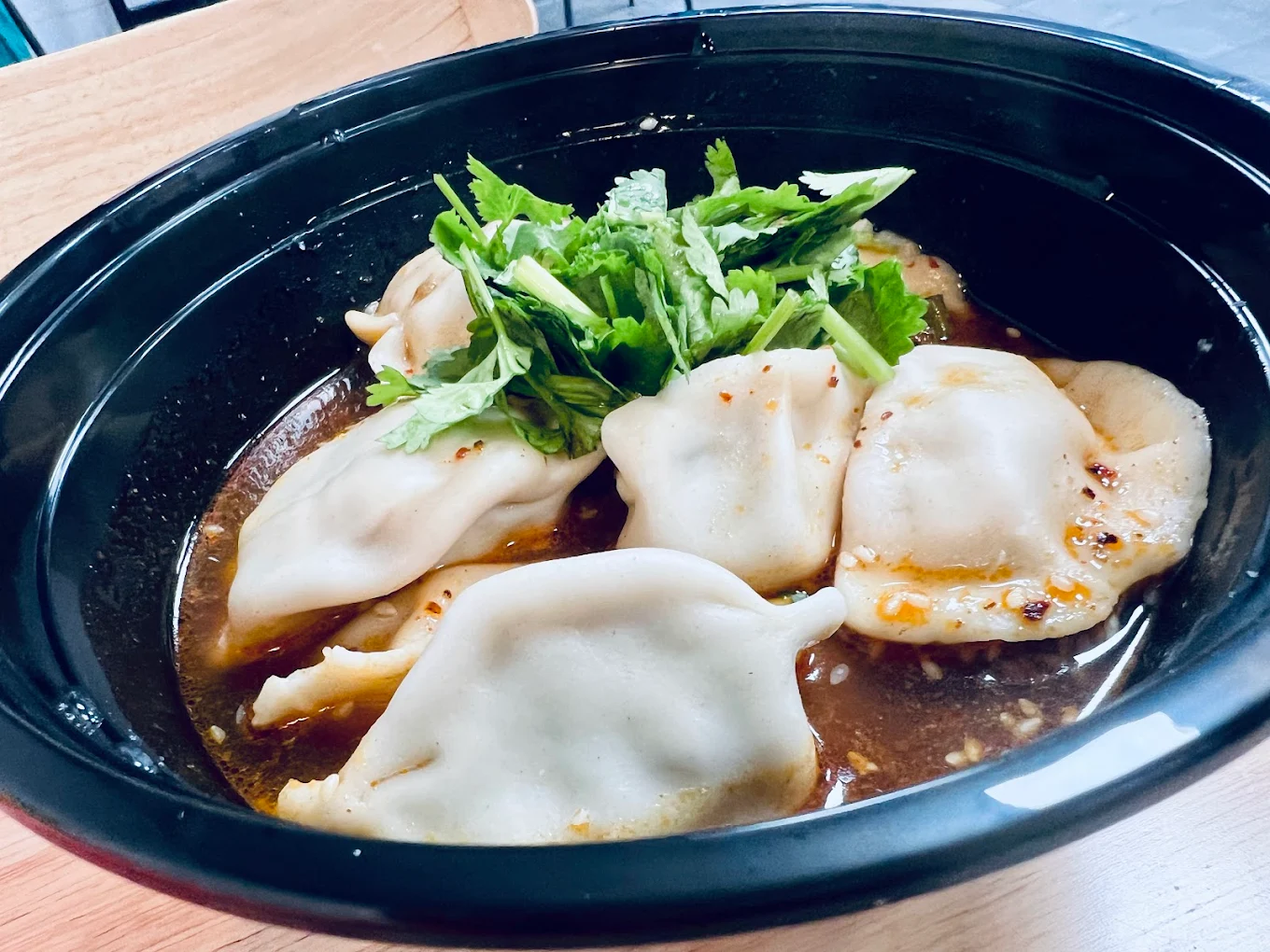 A close-up of a black bowl filled with dumplings in a savory broth, garnished with fresh cilantro on top. The dumplings appear soft and are partially submerged in the flavorful liquid.