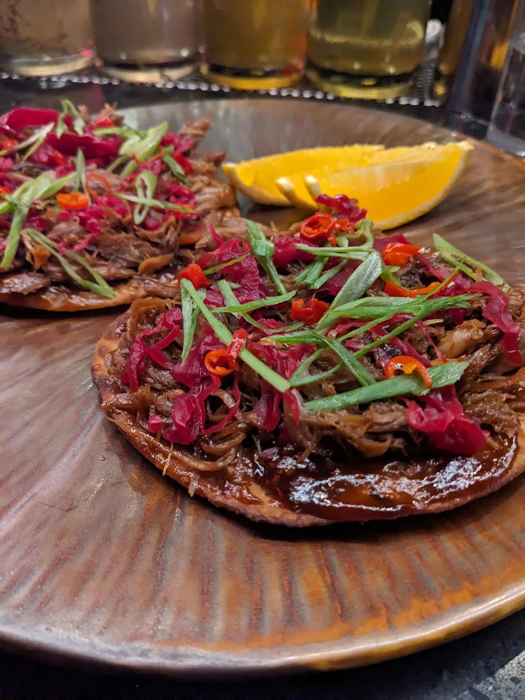 Two crispy tostadas topped with shredded meat, sliced green onions, red pickled vegetables, and sliced red chili peppers are served on a brown plate with orange wedges in the background.