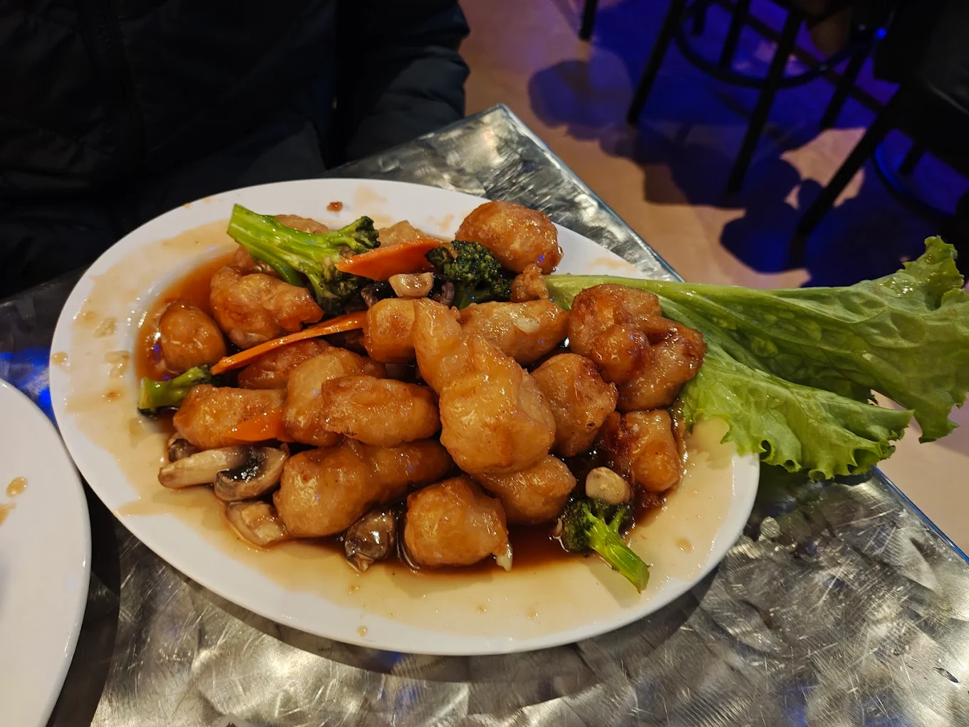 A white plate filled with battered and fried pieces of food in a brown sauce, garnished with mushrooms, broccoli, carrot strips, and a large green leaf, sits on a metallic table.