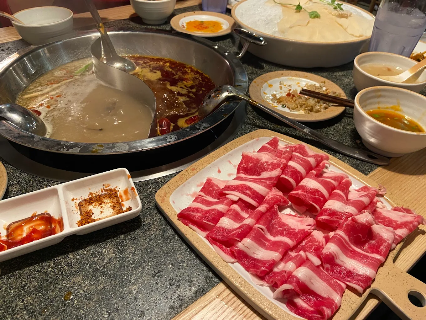 A hot pot meal with a divided pot of broth, thinly sliced raw beef on a tray, dipping sauces, and bowls with various ingredients set on a restaurant table.