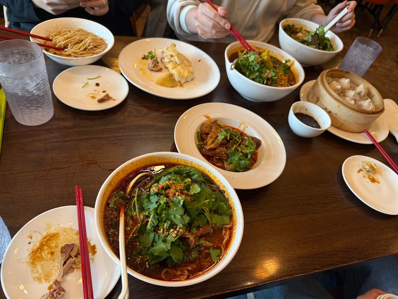 A table with bowls of noodle soup, plates of food, a basket of dumplings, and drinks. Three people are using chopsticks to eat, with various dishes spread out in a casual dining setting.