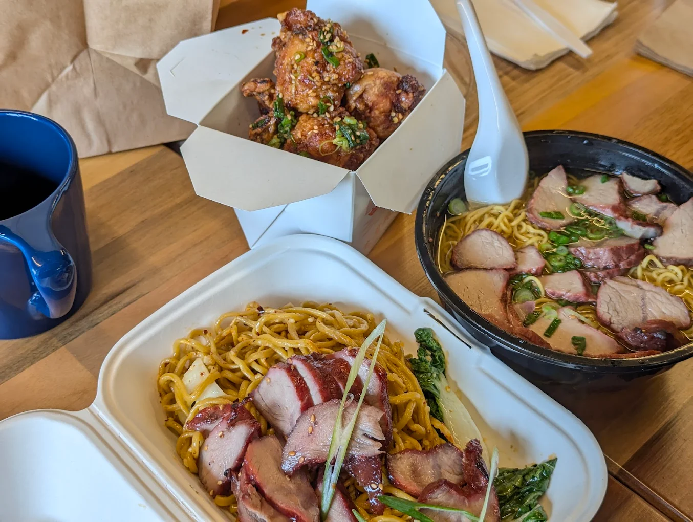 A table with takeout containers of Asian food: roasted pork over noodles, a bowl of ramen with sliced pork, and fried chicken wings topped with sesame seeds and scallions. A blue mug and wooden chopsticks are also visible.