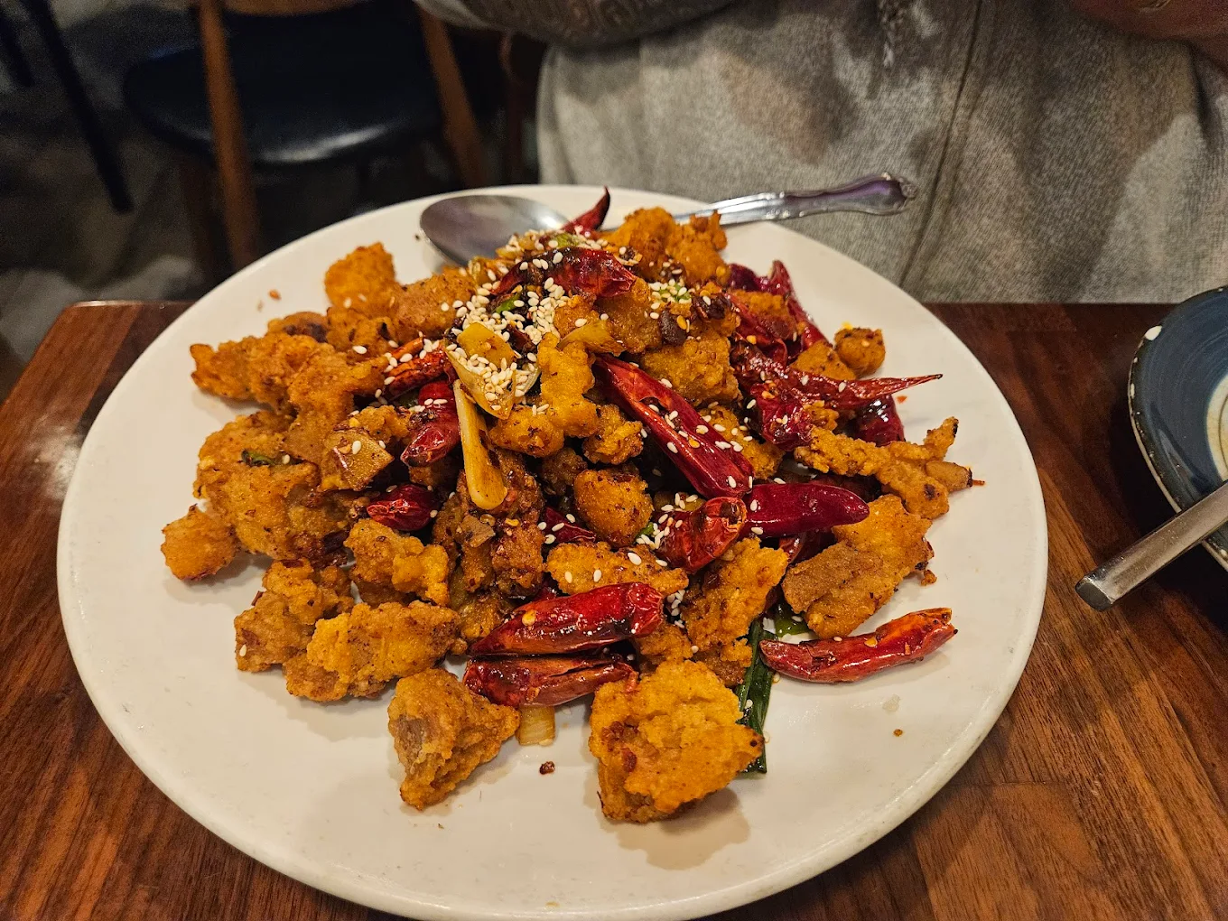 A plate of crispy fried chicken pieces topped with whole dried red chilies, garlic slices, and sprinkled with sesame seeds, served on a white plate with a spoon on the side.