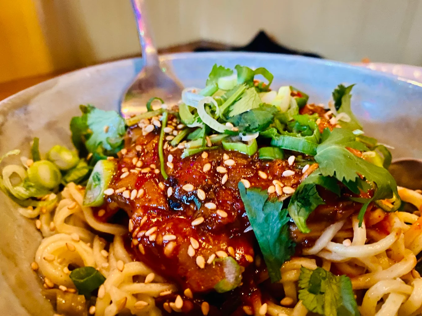 A close-up of a bowl of noodles topped with a spicy red sauce, chopped green onions, fresh cilantro, and sesame seeds, with a fork resting in the bowl.