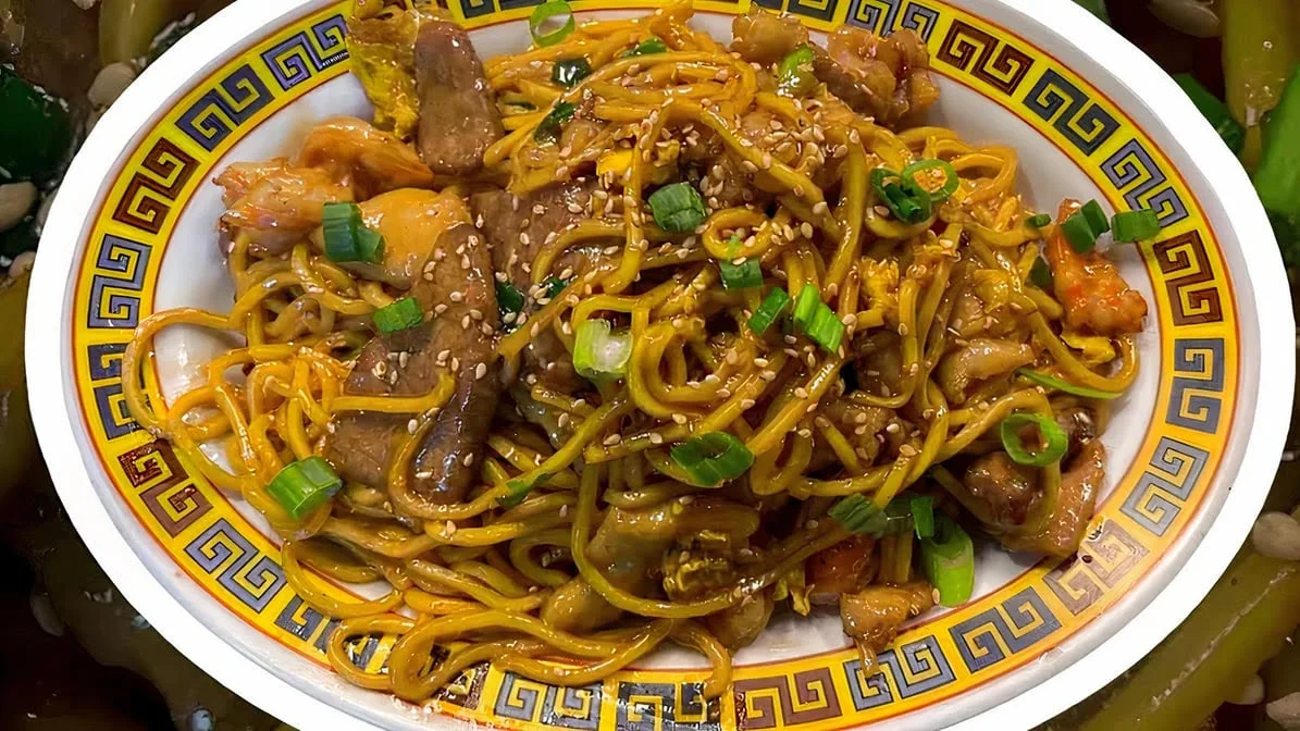 A plate of stir-fried noodles with chicken, vegetables, sliced green onions, and sesame seeds, served on a decorative plate with a blue and gold geometric border.