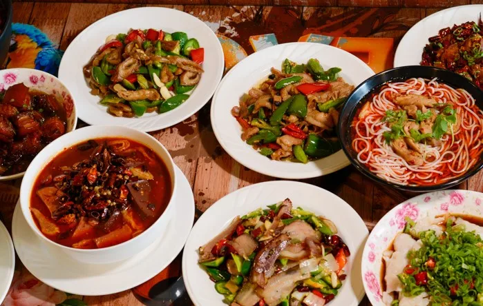 A table filled with various Chinese dishes, including plates of stir-fried vegetables and meats, bowls of noodle soup, and braised dishes, all colorful and richly garnished.