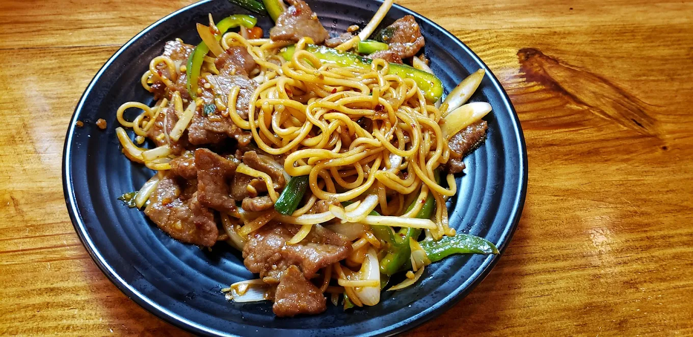 A dark plate of stir-fried noodles with beef strips, green bell peppers, onions, and bean sprouts, served on a wooden table.