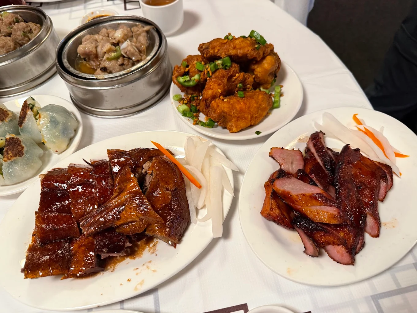 A table with plates of Chinese dishes, including roasted duck, char siu pork, fried chicken wings with green onions, and dim sum in metal steamers, set on a white tablecloth.