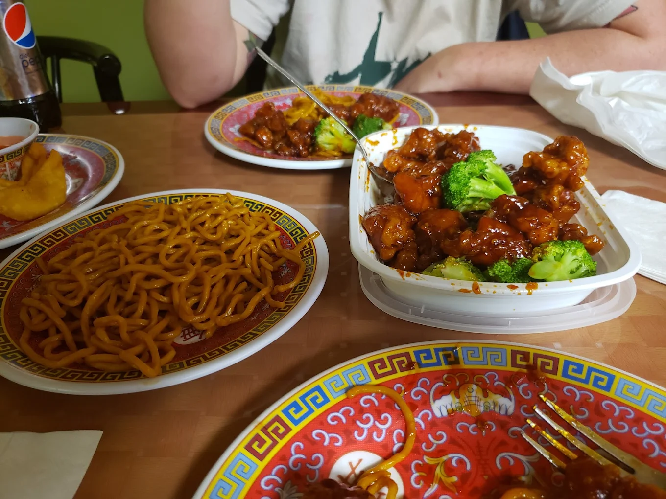 A table set with Chinese takeout food, including a container of saucy chicken with broccoli, a plate of noodles, and a colorful plate with food remnants. A person sits in the background with a can of Pepsi nearby.