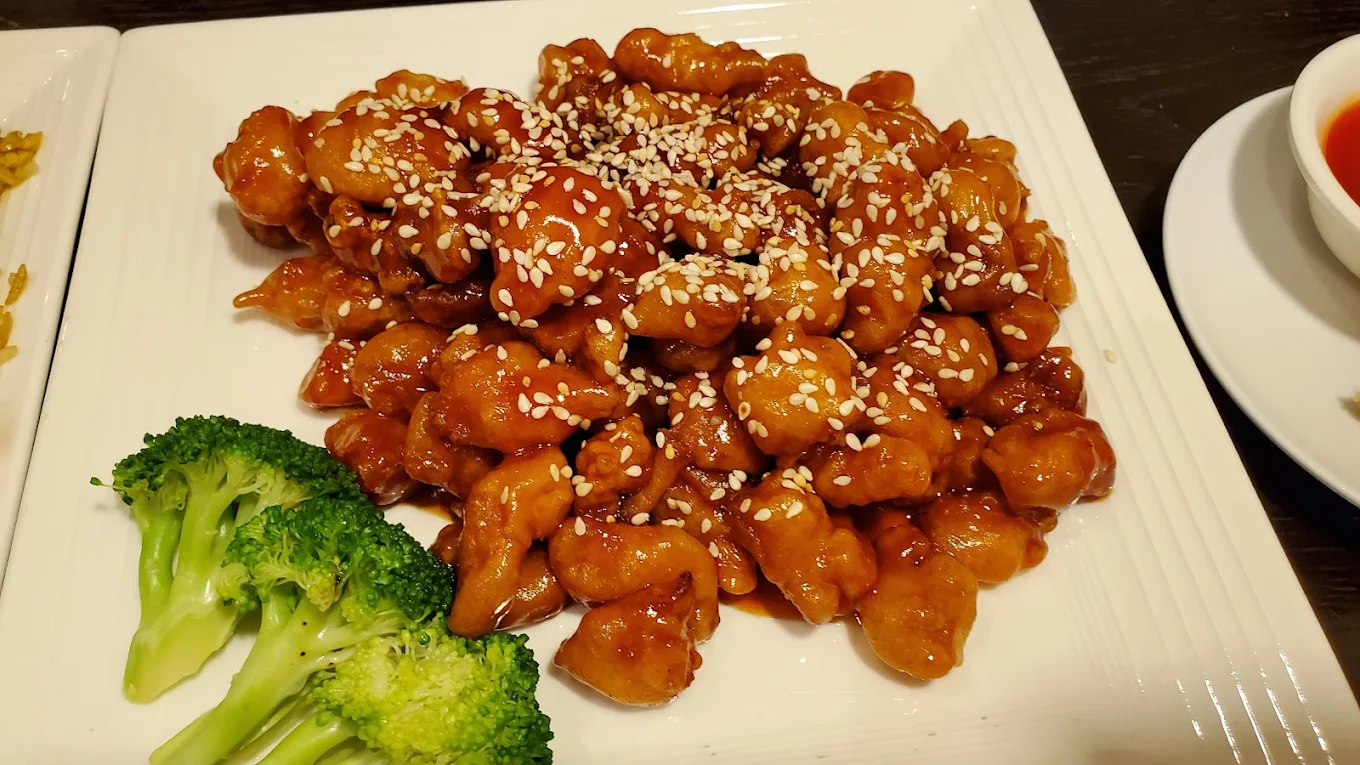 A plate of sesame chicken topped with sesame seeds, served with steamed broccoli on the side. The dish is glazed with a shiny, reddish-brown sauce.