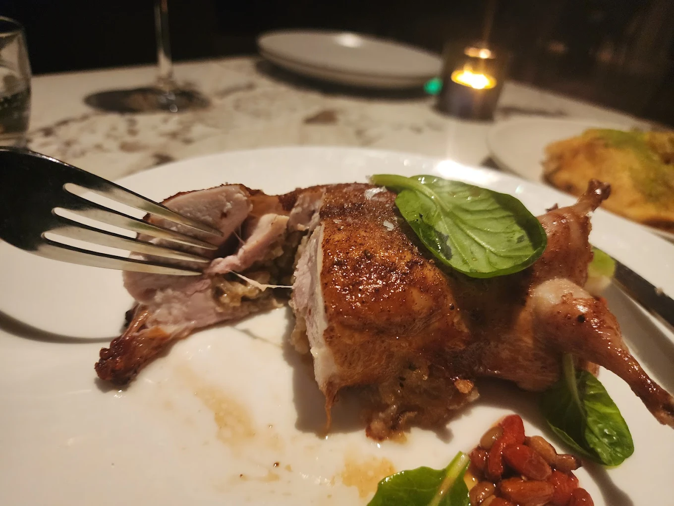 A close-up of a fork cutting into a roasted stuffed poultry dish on a white plate, garnished with basil leaves, with some vegetables on the side and a lit candle in the background.