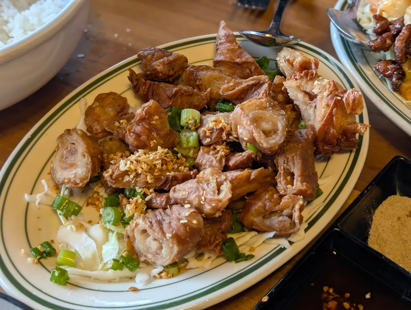 A plate of crispy fried pork intestines garnished with chopped green onions and fried garlic, served on a bed of shredded cabbage. Side dishes, rice, and dipping sauces are partially visible on the table.