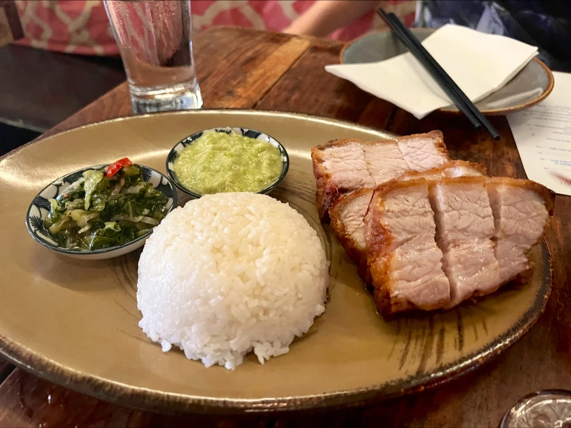 A plate with white rice, crispy pork belly slices, a bowl of green sauce, and a side of pickled vegetables. Black chopsticks rest on a napkin, and a glass of water sits on the wooden table.