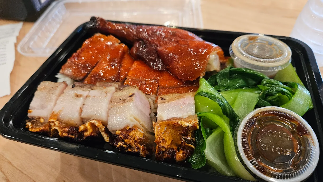 A takeout container with sliced roast duck, crispy pork belly, steamed bok choy, and two small containers of sauce, all served over white rice.