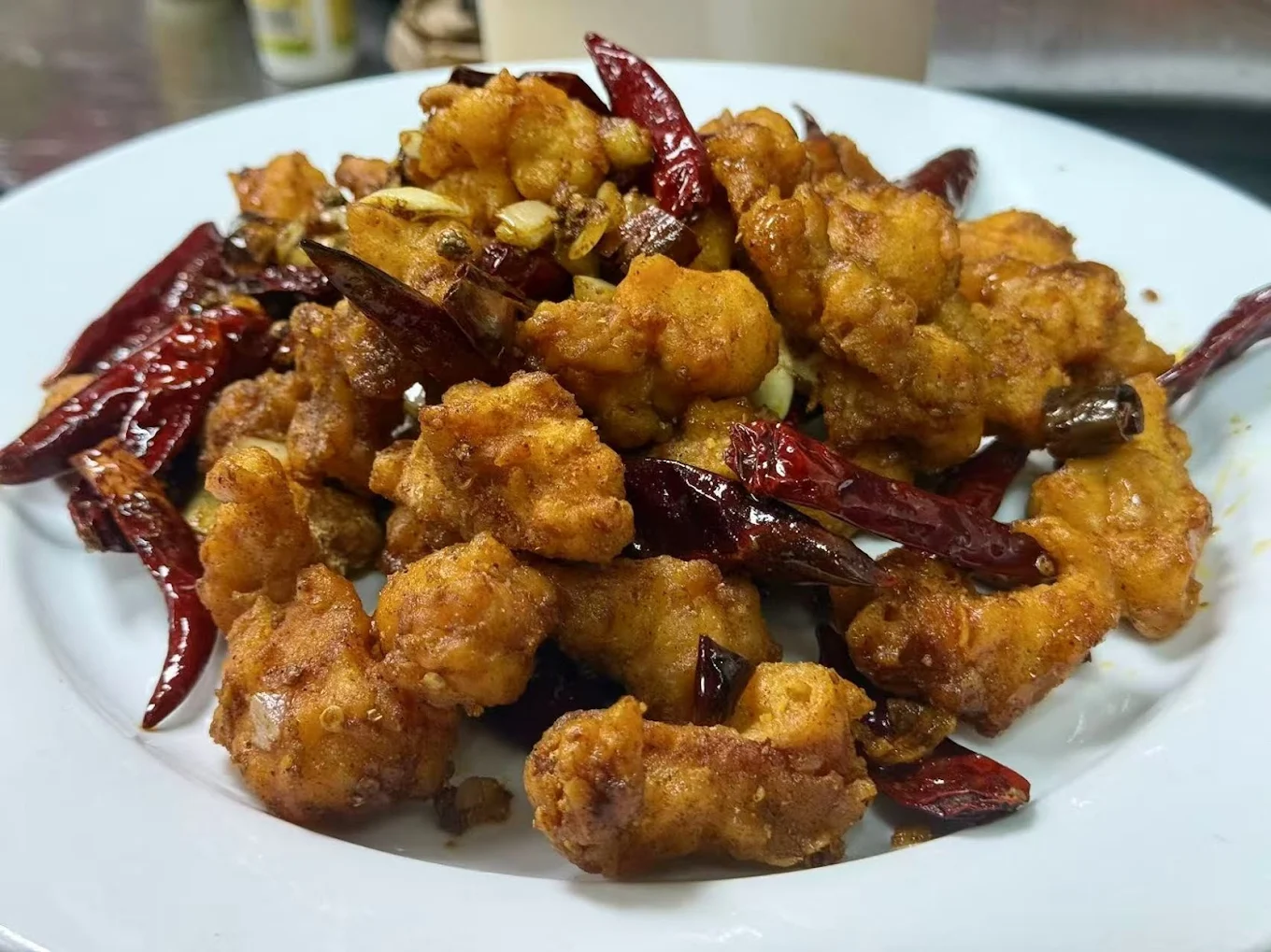 A plate of spicy, golden-brown fried chicken pieces garnished with whole dried red chilies and chopped garlic, served on a white dish.