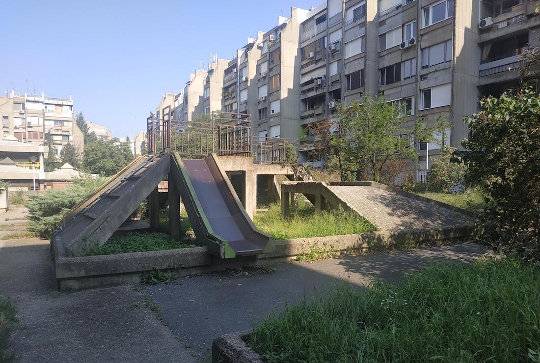 An old, concrete playground slide with metal sides stands in an overgrown grassy area between two rows of weathered apartment buildings on a sunny day.
