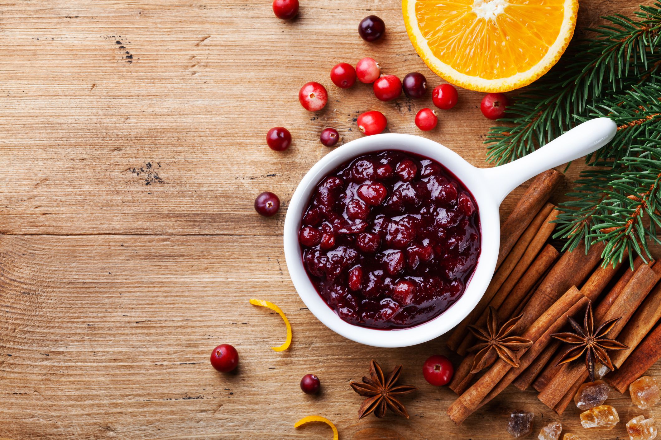 Cranberry sauce in ceramic saucepan with ingredients for cooking decorated with fir tree for Christmas or Thanksgiving day on rustic kitchen table. Top view with copy space.
