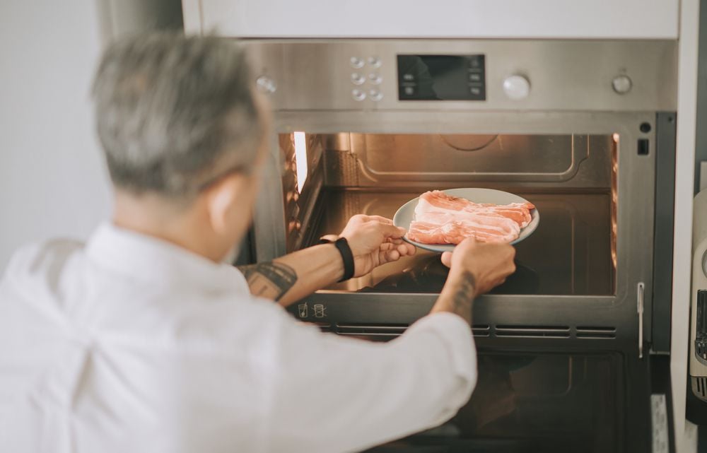 high angle view Asian chinese gray hair senior man with hair stubble defrosting pork meat with microwave preparing food cooking at home kitchen weekend