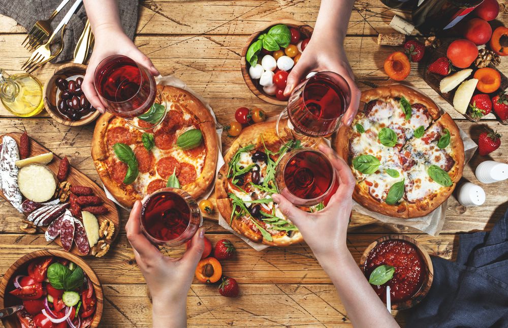 Pizza party with friends. Top view people holding glasses with red wine, rustic wooden table with hot pizzas, italian appetizers, salads, cheese, fruits and berries. Family lunch with fast food