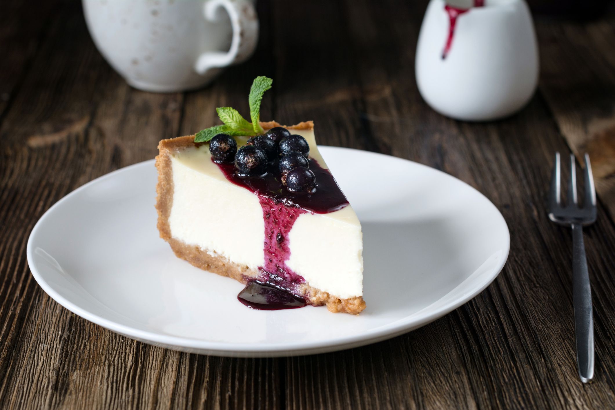 Piece of cheesecake with black currant and blueberry sauce on white plate on wooden table