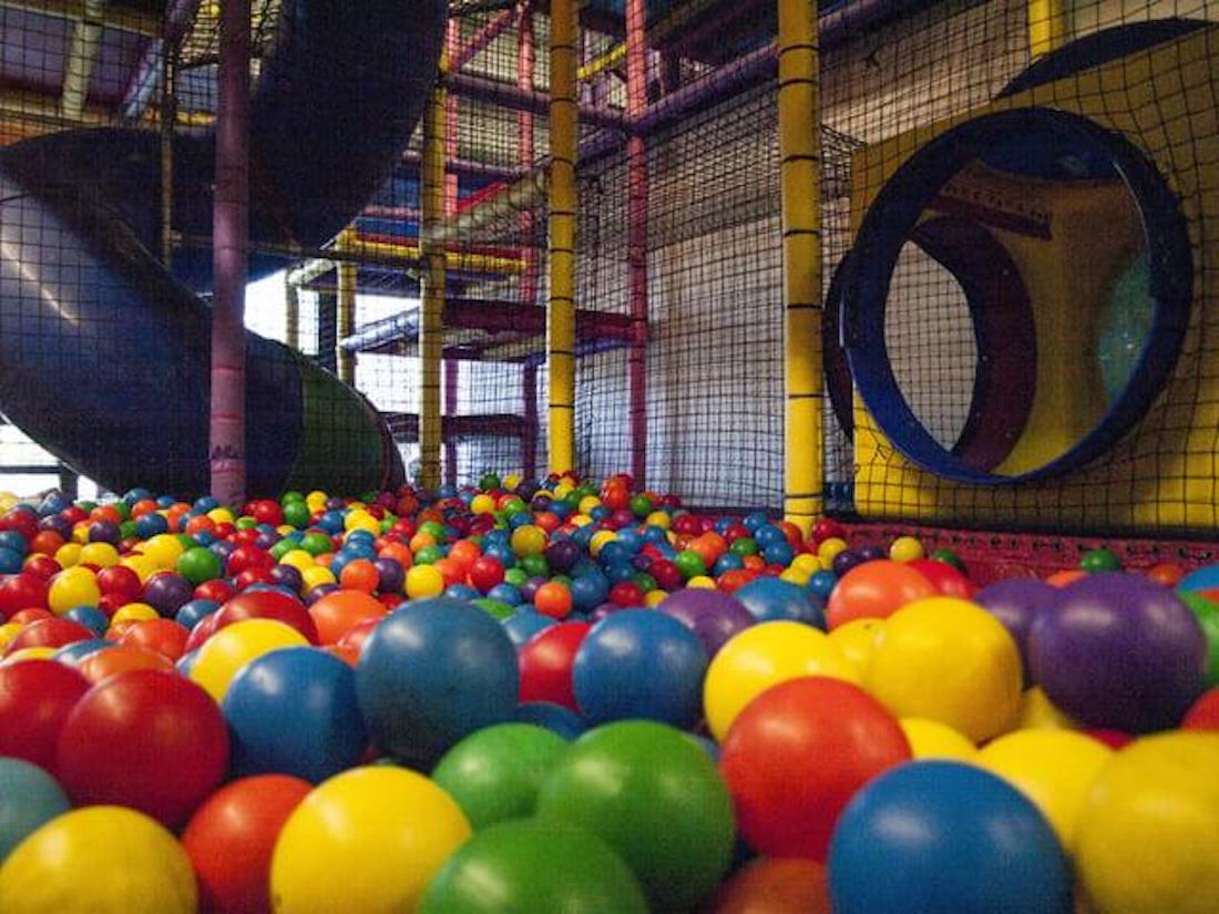 Indoor playground with a colorful ball pit, tunnel entrance, and slides surrounded by netted safety barriers. Multicolored plastic balls cover the floor.
